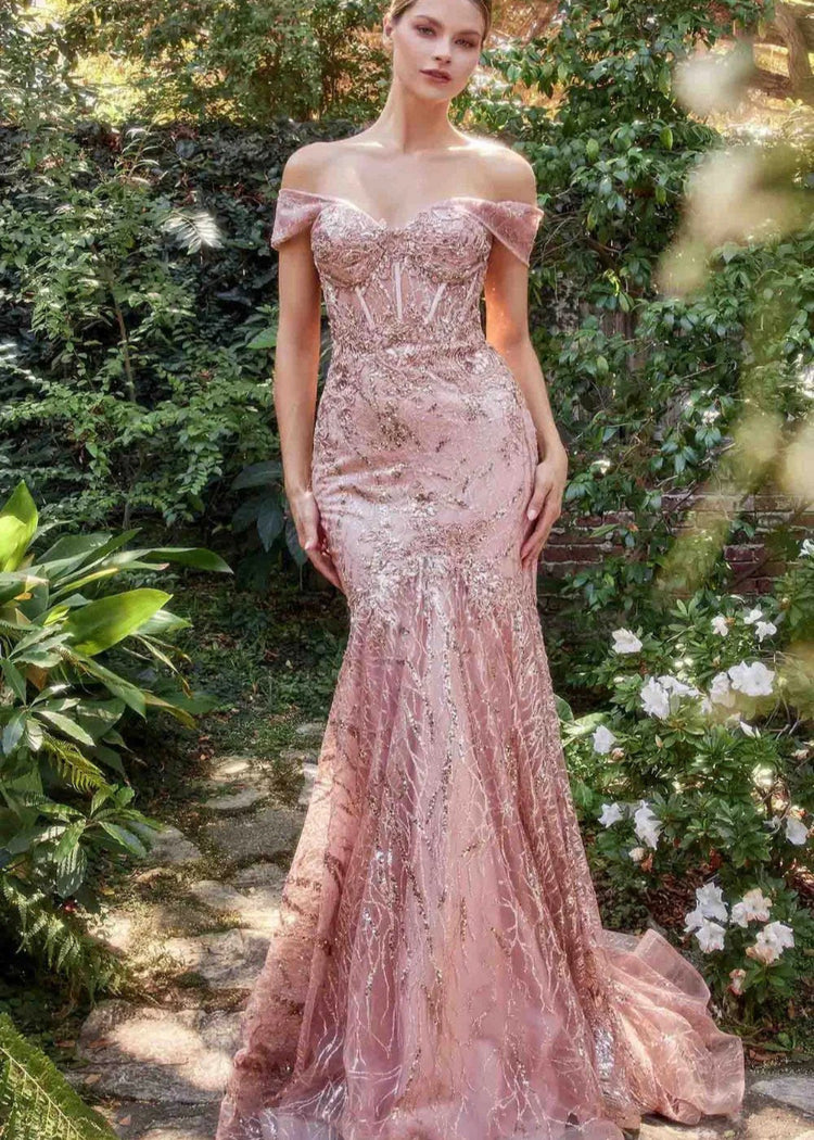 A model in a pink mermaid-style evening dress with glitter sequins, an off-the-shoulder neckline, and a sweep train, standing in a garden setting.