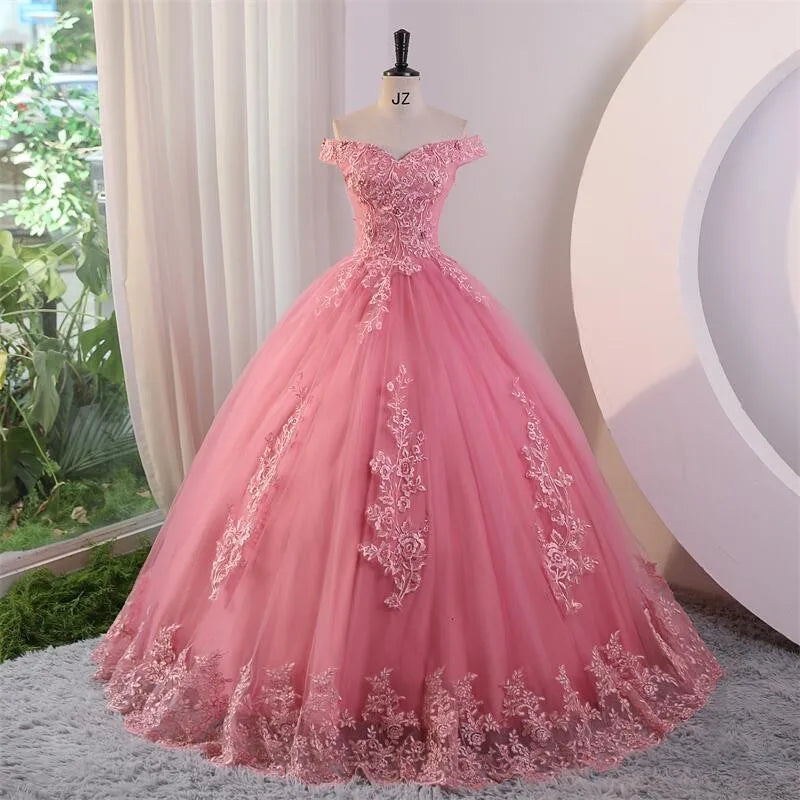A pink off-the-shoulder applique lace luxury ball gown with a floor-length skirt, displayed in a room with draped curtains.
